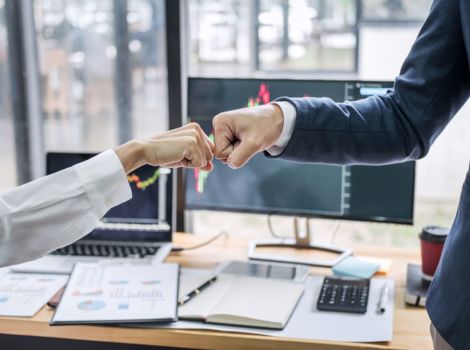 Zwei Personen stoßen vor einem Büro-Schreibtisch mit Ihren Fäusten an.