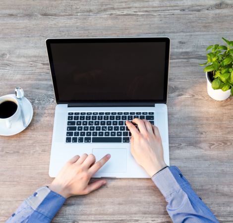 Ein Laptop steht auf einem Holztisch, daneben eine Tasse Kaffee und eine kleine Pflanze.