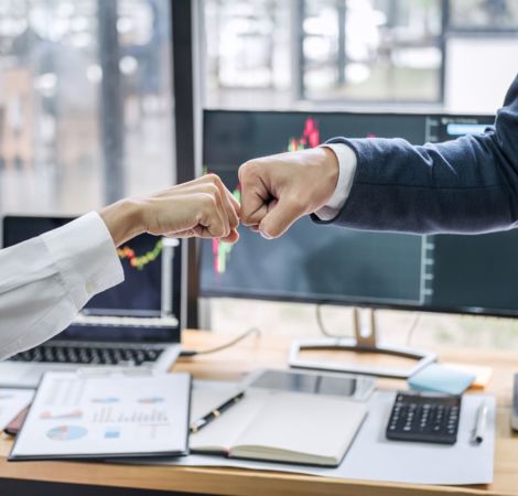 Zwei Personen stoßen vor einem Büro-Schreibtisch mit Ihren Fäusten an.