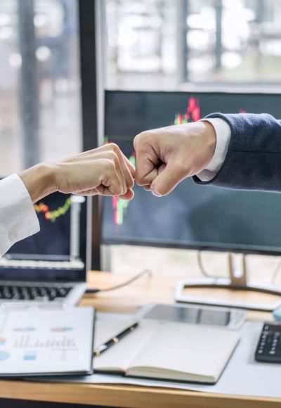 Zwei Personen stoßen vor einem Büro-Schreibtisch mit Ihren Fäusten an.
