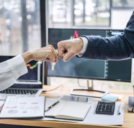 Zwei Personen stoßen vor einem Büro-Schreibtisch mit Ihren Fäusten an.